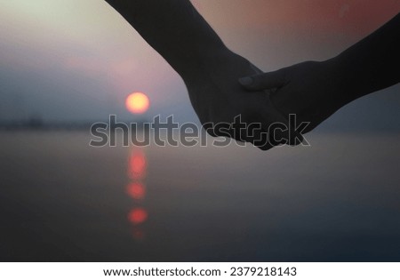 Close up of the hands of a romantic couple holding hands at sunset silhouetted against the colourful night sky with the orb of the sun visible below
