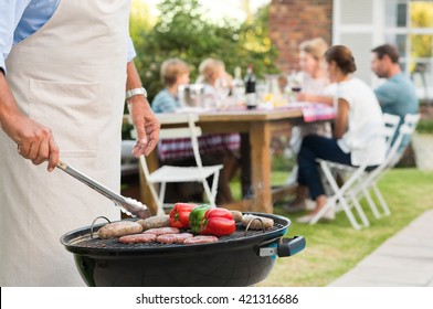 Close Up Hands Of A Senior Man Barbequing In The Garden With Family In Background. Grandfather Cooking On Grill. Man Cooking Hamburgers, Sausages And Peppers With Barbeque.