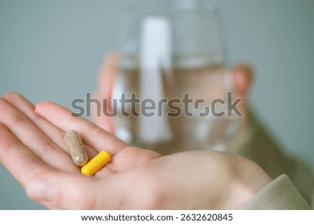 Close up hands showing with two capsules and a glass of water, illustrating a common practice of taking dietary supplements for better wellness in the morning. Health concept.