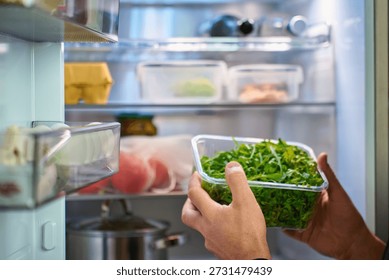 Close up of hands taking plastic container with fresh green arugula leaves from open refrigerator with food on shelves. Concept of healthy eating, home food storage and meal preparation