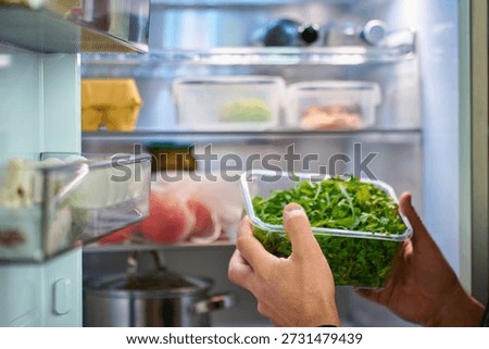 Close up of hands taking plastic container with fresh green arugula leaves from open refrigerator with food on shelves. Concept of healthy eating, home food storage and meal preparation
