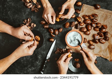 Close up of hands of three people peeling roasted chestnuts on black table background. Lifestyle family meal concept. Dark low key photo. Top view.