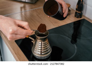 Close up of hands of unrecognizable man, putting full heaped tea spoon of natural ground coffee from round glossy tin in copper cezve on black electric cooker for brewing near spice grinder in kitchen - Powered by Shutterstock