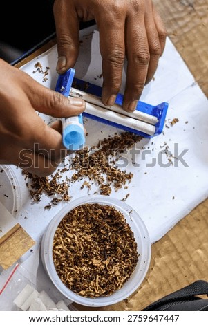 Close up of hands using a manual blue rolling machine to roll a cigarette with dried tobacco on a white table.