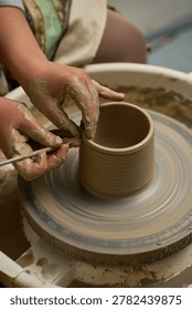 Close up of hands using a metal scraper to trim the rim of a wet clay pot on a wheel