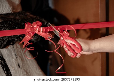 Close up of hands using pink scissors to cut a bright red ribbon with a decorative bow during a grand opening or inauguration ceremony.
