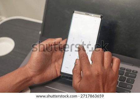 Close up of hands using a smartphone keypad with a laptop keyboard blurred in the background. Focus