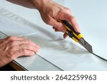 Close up of hands using utility knife cutting white vinyl adhesive film on glass table