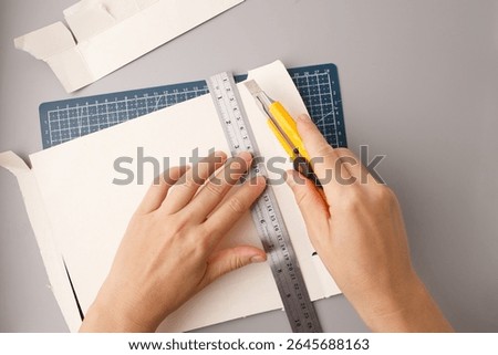 close up of hands using utility knife and metal ruler on cardboard with blue cutting mat perfect for crafting stores, educational tutorials, design studios, packaging prototypes, handmade art promo