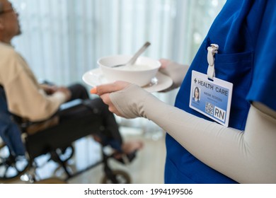 Close Up Hands Of Woman Nurse At Nursing Home Taking Care Of Disabled Senior Elderly Man On Wheelchair. Therapist Doctor Serving Food To Older Patient Male In House. Medical Insurance Service Concept.