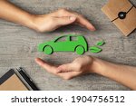 Close up of hands of woman protecting icon of green eco-friendly car over wooden table. Top view of woman hands protecting electric car with care, mobility. Car insurance and green automotive concept.