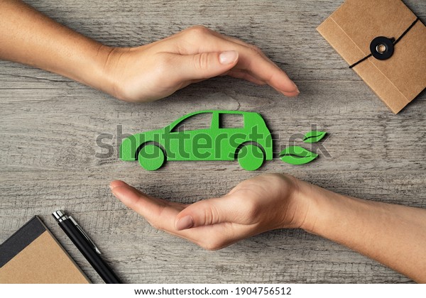 Close up of hands of woman protecting icon of\
green eco-friendly car over wooden table. Top view of woman hands\
protecting electric car with care, mobility. Car insurance and\
green automotive concept.