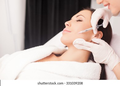 Close up of hands of young cosmetologist injecting botox in female face. She is standing and smiling. The woman is closed her eyes with relaxation