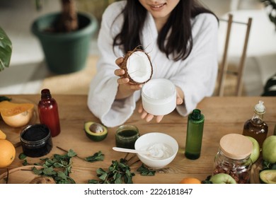 Close up hands of young woman holding half of coconut while making homemade cream for healthy skin. A lot of ingredients for home made cosmetics lying on wooden table. - Shutterstock ID 2226181847