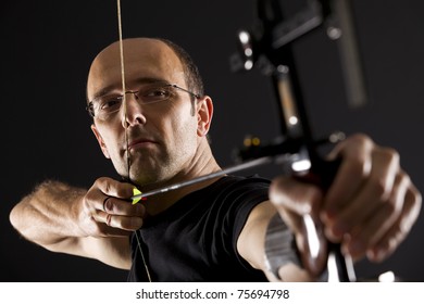 Close Up Of Handsome Archer In Black On Black Background Aiming With Bow And Arrow, Side View With Focus On Eyes.