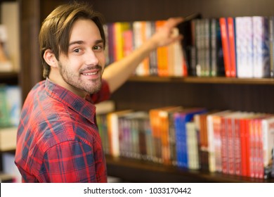 Close Up Of A Handsome Bearded Young Man Smiling To The Camera Over His Shoulder While Picking A Book From The Shelf At The Library Or Bookstore Copyspace Lifestyle Student Learner College Campus.