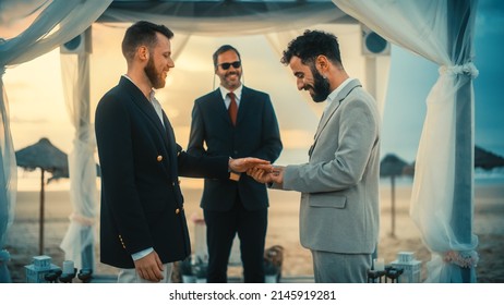 Close Up Of Handsome Gay Couple Exchange Rings And Kiss At A Beach Wedding Ceremony Venue At Sunset. Two Happy Men In Love Share Their Vows And Get Married. LGBTQ Relationship Goals.