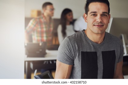 Close Up Of Handsome Male Worker Wearing Gray Short Sleeve Shirt In Small Office With Other Employees Behind Him