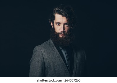 Close Up Handsome Young Man With Long Goatee Beard, Wearing An Elegant Gray Business Coat, Looking At Camera On A Black Background.