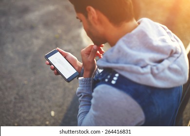 Close up handsome young man using smart phone while standing outdoors at sunny evening, rear view shot hipster man holding cell phone while looking to some fashion website on mobile screen, flare