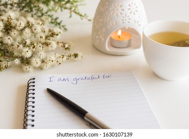 Close up of handwritten text "I am grateful for..." in foreground with notebook, pen,  cup of tea, flowers and oil burner in soft focus (deliberate angle)
