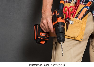 Close up of handyman holding a drill machine with tool belt around waist. Detail of artisan hand holding electric drill isolated over grey background. Hand of bricklayer holding carpentry accessories.