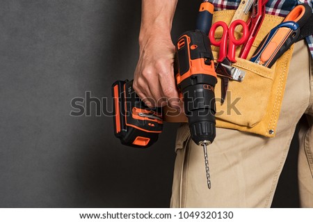 Close up of handyman holding a drill machine with tool belt around waist. Detail of artisan hand holding electric drill isolated over grey background. Hand of bricklayer holding carpentry accessories.