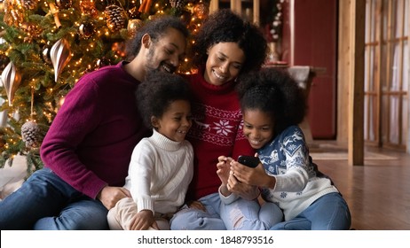 Close Up Happy African American Family Wearing Sweaters Using Smartphone, Sitting On Warm Floor Near Christmas Tree At Home, Smiling Mother And Father With Kids Spending Winter Holidays Together