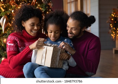 Close Up Happy African American Family Unpacking Christmas Gift, Smiling Mother And Father With Adorable Little Daughter Unwrapping Present Box, Sitting Near Festive Tree At Home, Winter Holiday