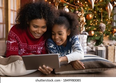 Close Up Happy African American Woman With Daughter Using Tablet Together, Lying On Soft Pillows Near Christmas Tree At Home, Happy Family Shopping Online, Choosing Gifts, Enjoying Winter Holiday