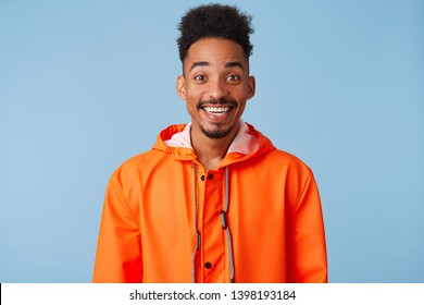 Close up of happy amazed young african american dark skinned man, feels great, wears in orange rain coat, broadly smiles isolated over blue background.