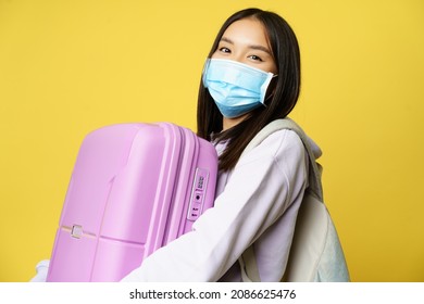Close up of happy asian girl tourist in medical face mask, holding pink cute suitcase, going on vacation, travelling during pandemic, yellow background