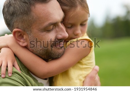 Close up of happy dad holding his lovely little daughter, smiling, standing with eyes closed while spending time together outdoors on a warm day