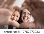 Close up happy faces view though joined fingers showing love symbol, granny and little granddaughter smile, revealing white teeth, advertising dental clinic service, medical insurance cover for family