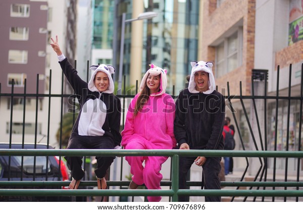 Close up of a happy group of friends, waiting the\
public transportation and wearing different costumes, one woman\
wearing a pink unicorn costume, other woman a panda costume and the\
man wearing a cat