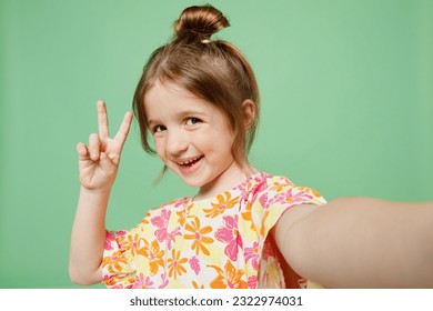 Close up happy little child kid girl 6-7 years old wear casual clothes have fun do selfie shot pov on mobile cell phone show v-sign isolated on plain green background. Mother's Day love family concept