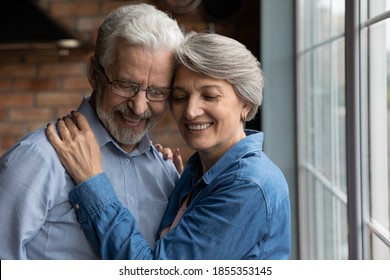 Close up happy mature couple hugging with closed eyes, standing in kitchen at home, elderly family enjoying tender moment, smiling senior woman and man wearing glasses cuddling, feeling love - Powered by Shutterstock