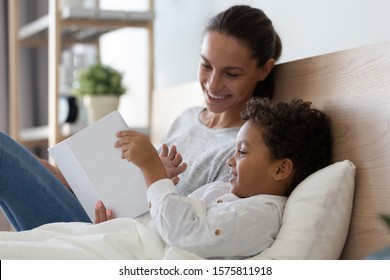 Close up happy mixed race family lying on pillows in bed, holding book, watching pictures together. Smiling young mommy reading fairytale to little cute multiracial preschool son at modern bedroom. - Powered by Shutterstock