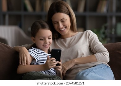 Close Up Happy Mother With Little Daughter Kid Using Smartphone Sitting On Couch At Home, Smiling Caring Mom And Adorable Girl Child Looking At Device Screen, Chatting Online, Watching Cartoons