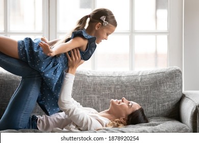 Close Up Happy Mother Playing Active Game With Little Daughter, Lying On Couch At Home, Smiling Mum Holding Lifting Adorable Girl Child With Hands Outstretched Pretending Flying, Enjoying Weekend