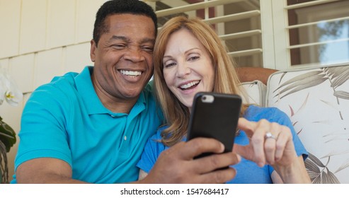 Close Up Of Happy Older Couple Sitting On Porch And Using Smartphone Together. African American And Caucasian Husband And Wife Looking At Cell Phone On Sunny Day Outside Home