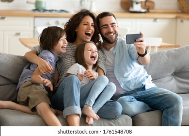 Close Up Happy Parents With Little Children Using Smartphone Together Sitting On Couch At Home. Smiling Father Holding Phone Taking Selfie With Kids. Family Watching Video Having Fun With Cellphone.