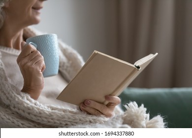 Close Up Happy Smiling Elderly Senior Woman Drinking Hot Tea Coffee, Reading Favorite Bestseller Novel Poetry In Paper Book, Enjoying Cozy Lazy Weekend Holiday Time, Relaxing Alone On Sofa At Home.