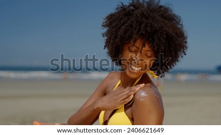 Close up of a happy smiling young black woman is applying a sunscreen or sun tanning lotion on a shoulder to take care of her skin on a seaside beach during holidays vacation.
