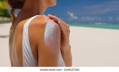 Close up of a happy smiling young woman is applying a sunscreen or sun tanning lotion on a shoulder to take care of her skin on a seaside beach during holidays vacation.