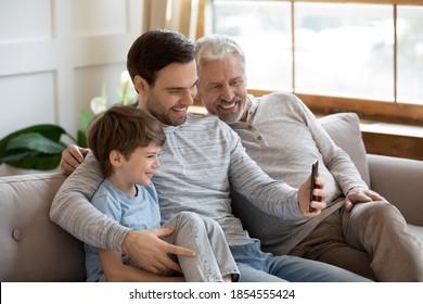 Close up happy three generations of men using smartphone together, having fun with gadget, overjoyed little boy with mature grandfather and father looking at phone screen, sitting on couch at home - Powered by Shutterstock