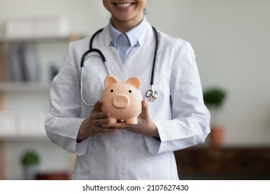 Close up happy young Indian female GP doctor holding piggy bank in hands, advertising collecting costs for medical insurance or charity donation help in clinic office, healthcare and finances concept.