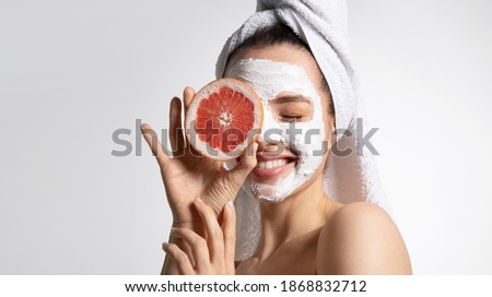 Close up of happy young playful teen girl in moisturizing mask and towel holds grapefruit covers eye. Advertising poster of facial eco-friendly skincare products. Morning beauty routine