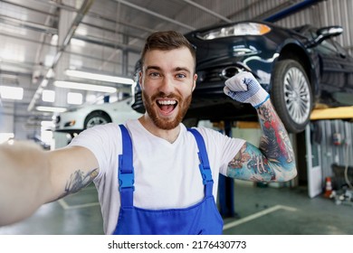 Close up happy young professional technician mechanic man in blue overalls t-shirt do selfie shot pov mobile phone stand near car lift do winner gesture work in vehicle repair shop workshop indoors - Powered by Shutterstock