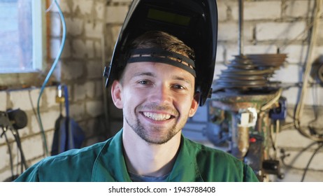 Close up of happy young repairman opens protective mask after finishing his job. Portrait of mechanic in workwear smiling and looking into camera. Welder with beard working at garage. Slow motion.